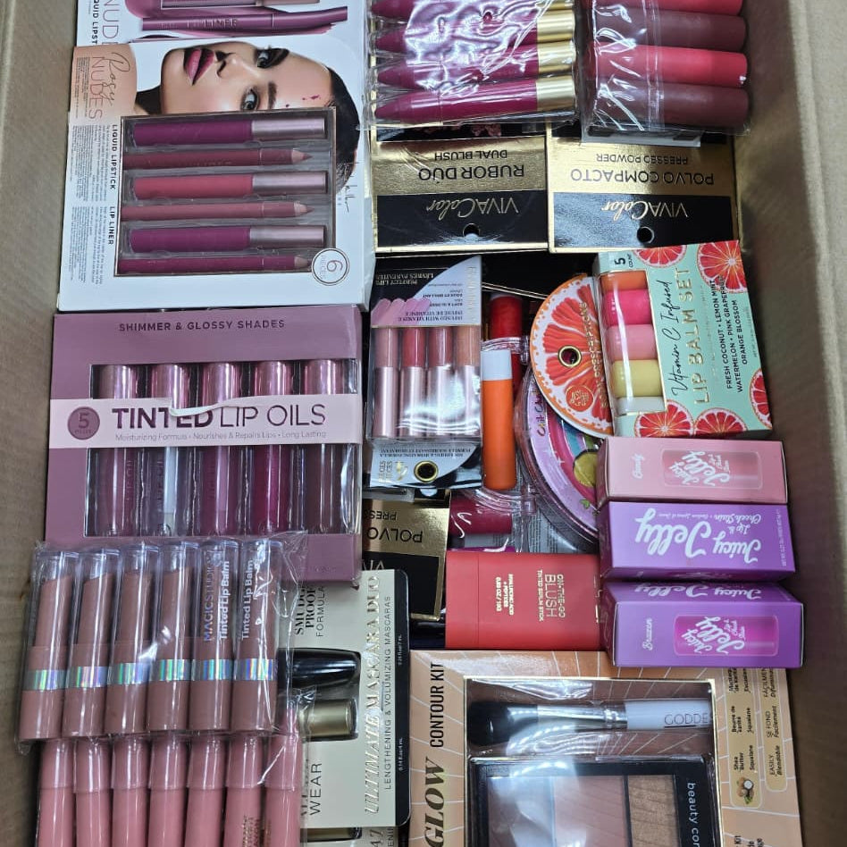 A cardboard box filled with multiple makeup items and beauty products, including lip glosses, lip oils, compacts, palettes, and brushes, photographed from above.