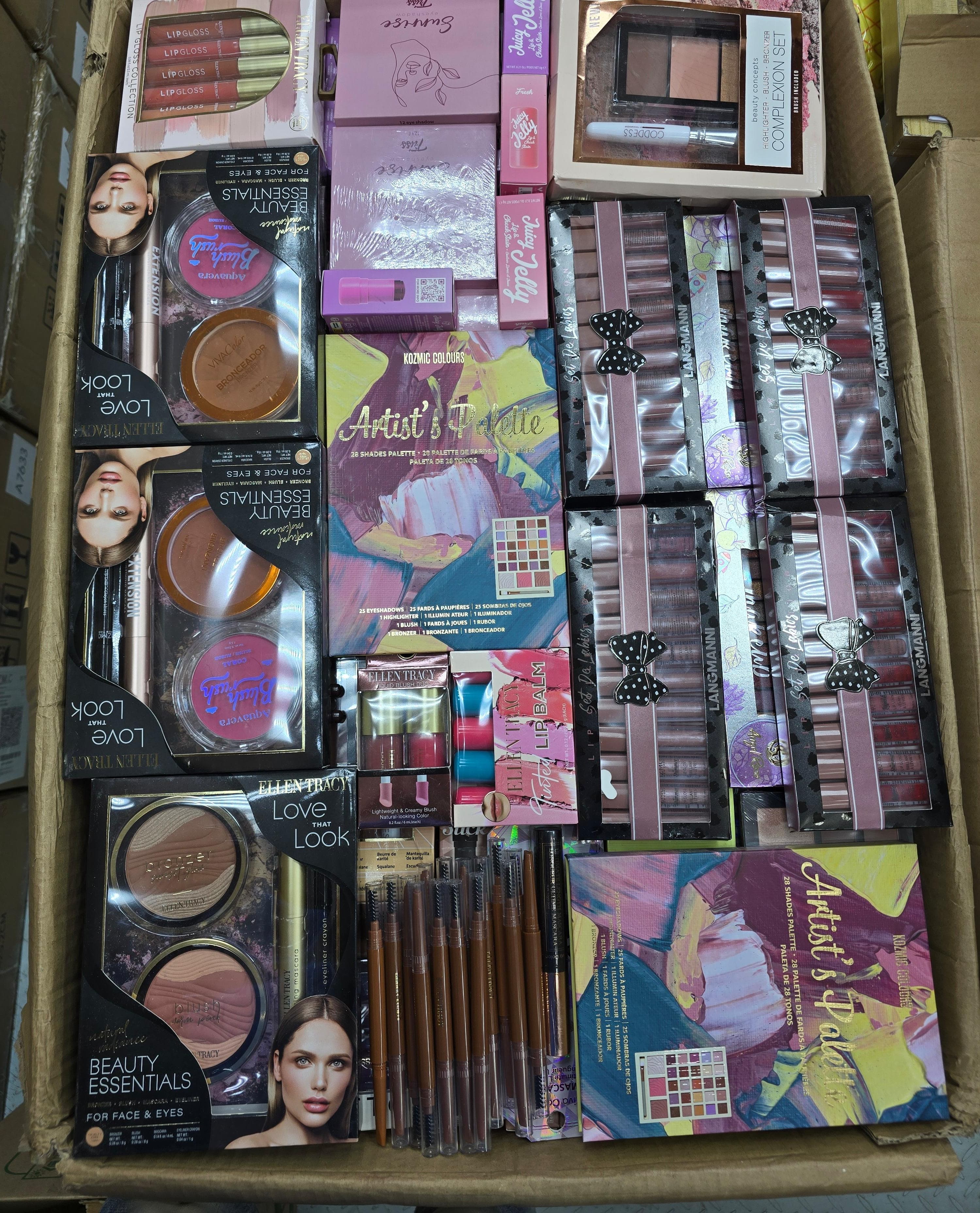 A bulk lot of assorted makeup products packed in a cardboard box, including eyeshadow palettes, blushes, lipsticks, brushes, and palettes from brands like Beauty Creations and Artist's Palette.